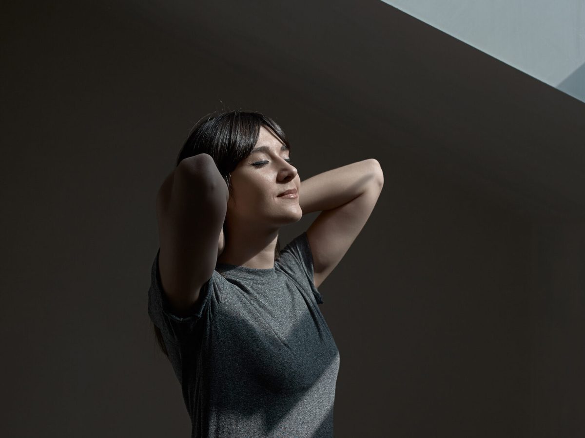 Smiling girl with closed eyes breathing fresh air standing near window in shadows with light reflected on her face, holding her hands behind her neck.