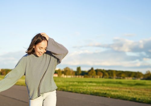 Vertical portrait of happy asian girl enjoying skateboard fun day out. Smiling korean skater on longboard, riding along empty street on sunny day.